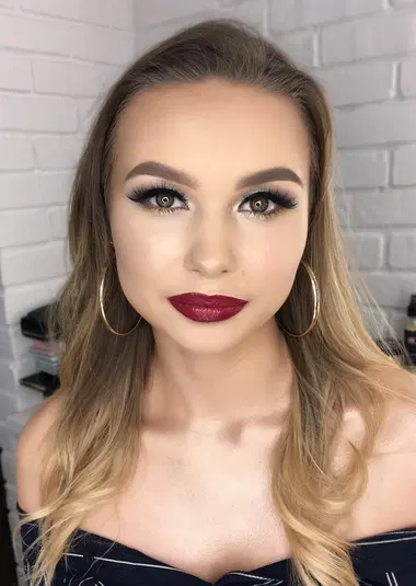  Młoda kobieta z makijażem wieczorowym, podkreślone oczy i czerwone usta, nosząca złote kolczyki, blond włosy, sesja w studio z białą ceglaną ścianą w tle. 
