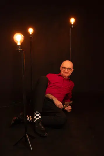  Osoba w czerwonej koszuli grająca na harmonijce ustnej, otoczona trzema stojącymi lampami, na ciemnym tle. 