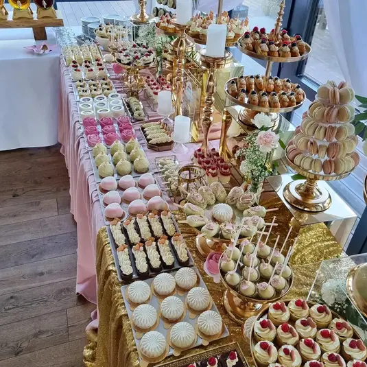  Stół pełen eleganckich słodkości na weselny candy bar, z różnorodnymi ciastami, różowymi makaronikami, deserami w pucharkach i ozdobnymi babeczkami na złotych tacach, idealny na przyjęcie lub uroczystość. 