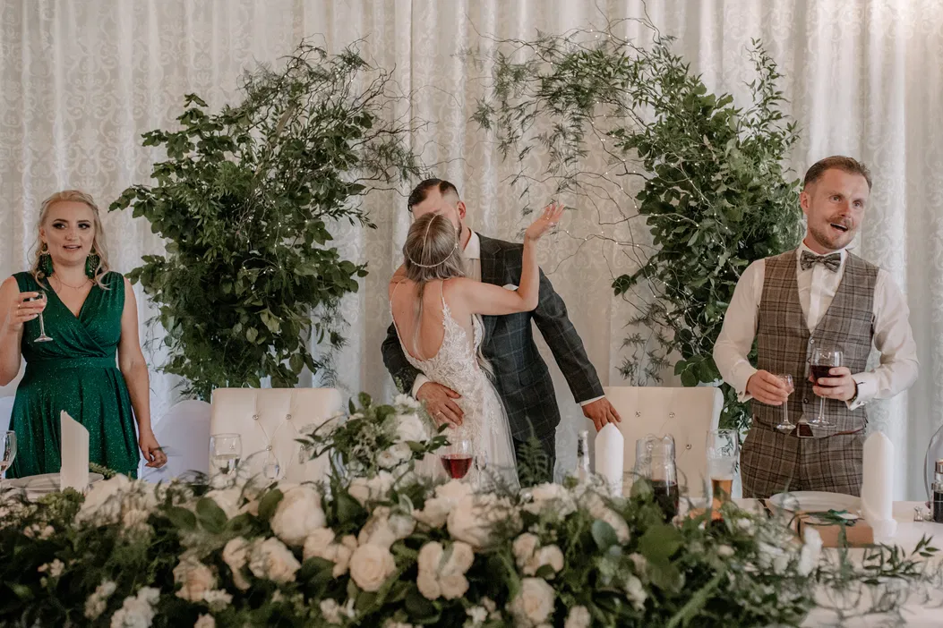 Para młoda całująca się na weselu, otoczona zielonymi dekoracjami i świadkami w eleganckich strojach.
