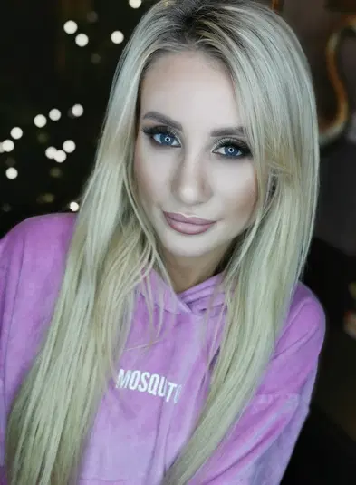  Blondynka w różowej bluzie Mosquito, z rozświetlonym makijażem, na ciemnym tle z efektem bokeh. 