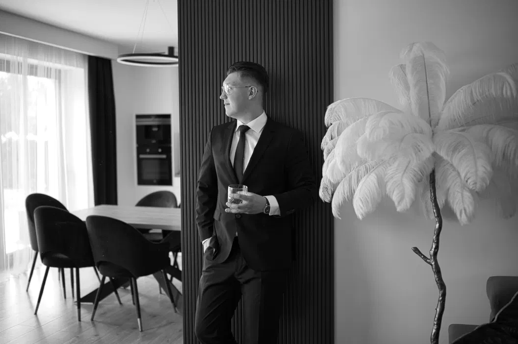 Mężczyzna w garniturze z drinkiem w ręku stojący w nowoczesnym salonie z eleganckimi meblami i dekoracyjną rośliną.