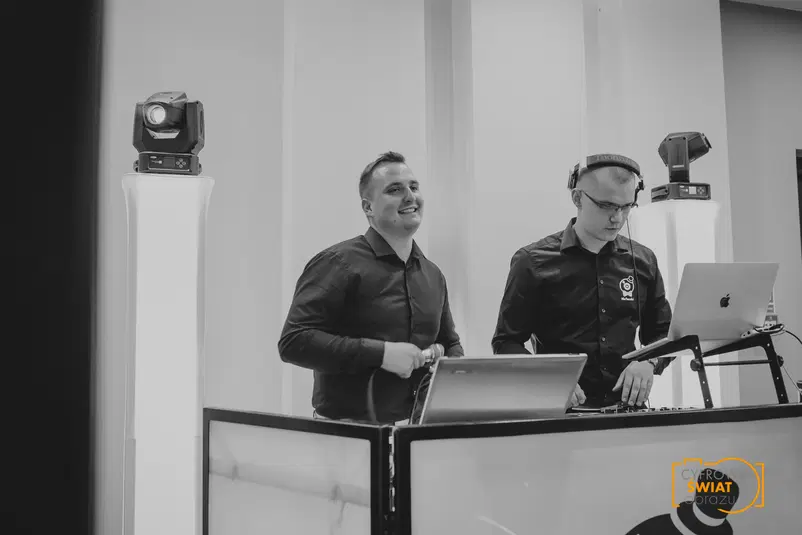  DJ-e na imprezie; dwóch mężczyzn przy konsoli DJ-skiej, jeden uśmiechnięty, drugi z słuchawkami, laptop; czarno-biała fotografia evenementowa. 