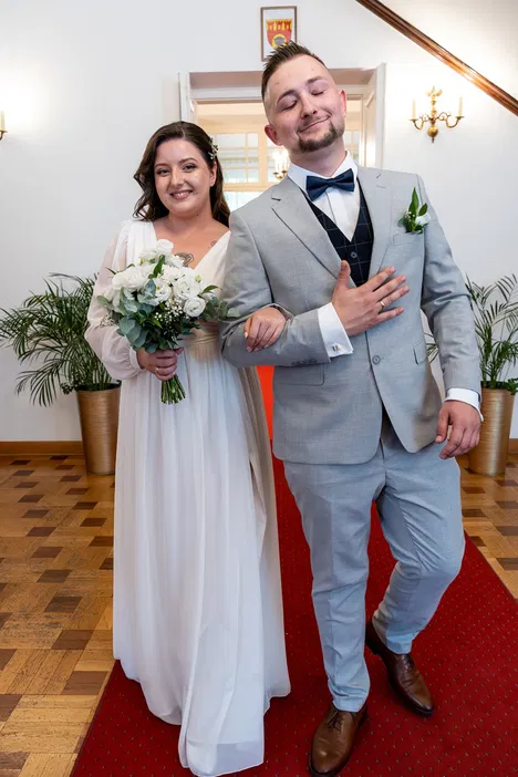 Młoda para w eleganckich strojach ślubnych, kobieta w białej sukni z bukietem kwiatów, mężczyzna w szarym garniturze z muszką, na czerwonym dywanie, w eleganckim wnętrzu.