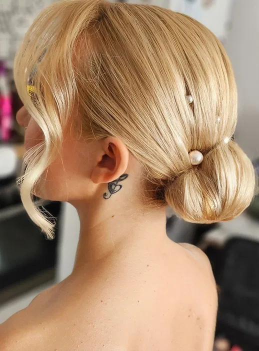 Blond włosy upięte w elegancki kok z perłowymi ozdobami, widoczny tatuaż za uchem.