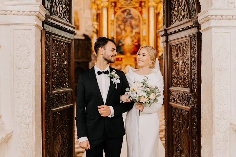  Para młoda wychodząca z kościoła po ceremonii ślubnej, elegancko ubrany pan młody i panna młoda w białej sukni z bukietem kwiatów, szczęśliwi i uśmiechnięci. 