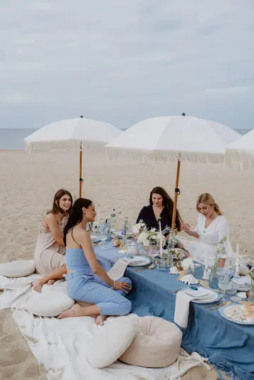  Cztery kobiety przy elegancko nakrytym stole na plaży, pod parasolami. 