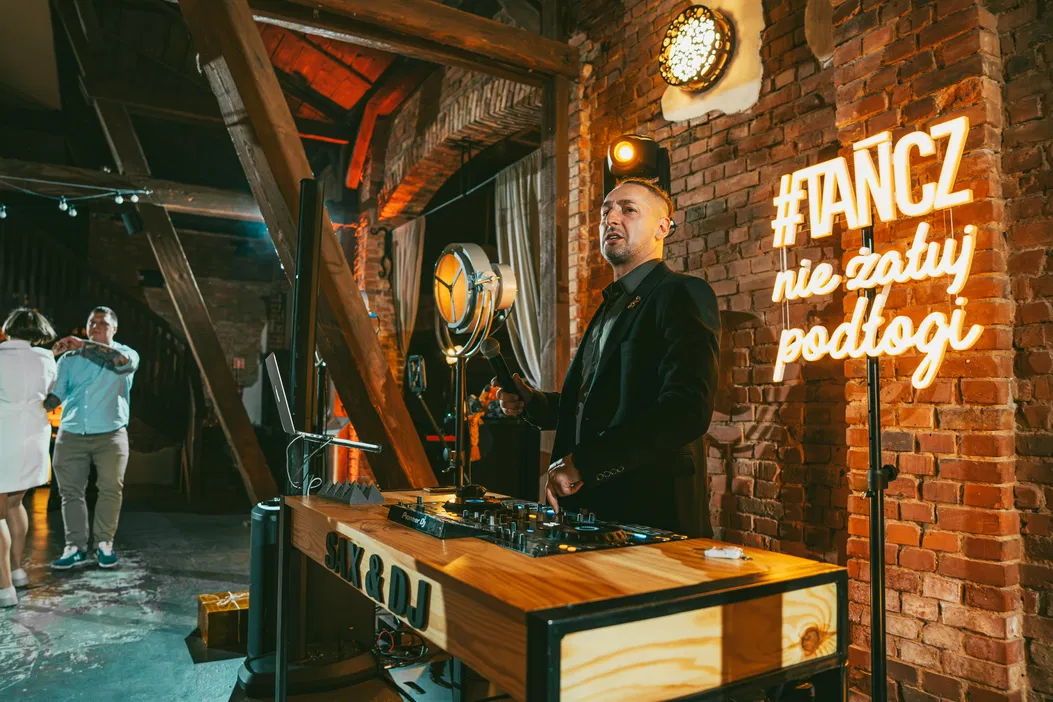DJ grający muzykę na imprezie w klimatycznym wnętrzu z neonowym napisem Tańcz, nie zatuj podłogi.
