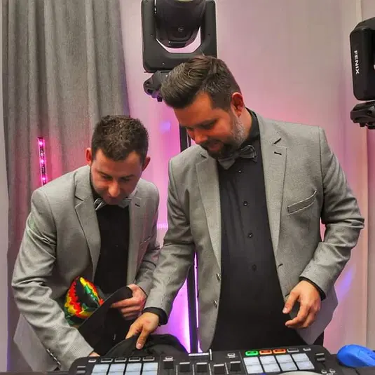  Dwóch mężczyzn w eleganckich garniturach przy konsoli DJ-skiej na przyjęciu. 