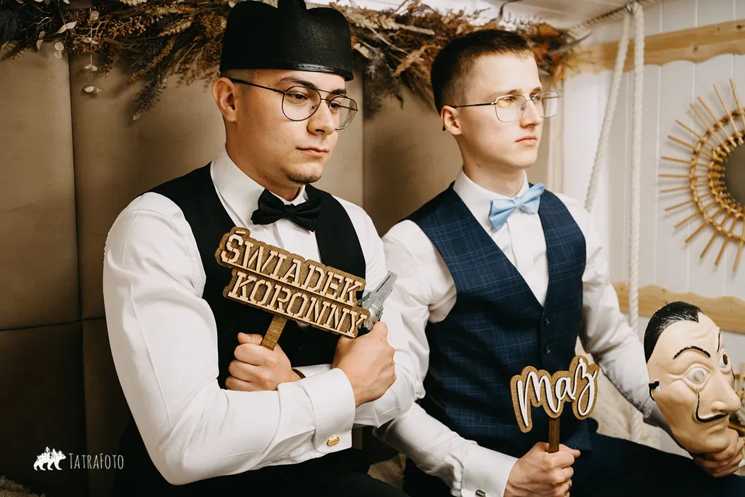 Dwóch młodych mężczyzn w eleganckich strojach na sesji zdjęciowej, jeden z podpisem Świadek Koronny, drugi z napisem Mąż, trzymając maskę.