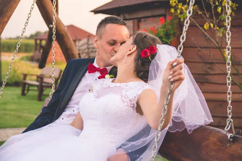  Para młoda na huśtawce, romantyczny pocałunek, wesele na świeżym powietrzu, panna młoda w białej sukni, pan młody w garniturze z muchą, dekoracje kwiatowe. 