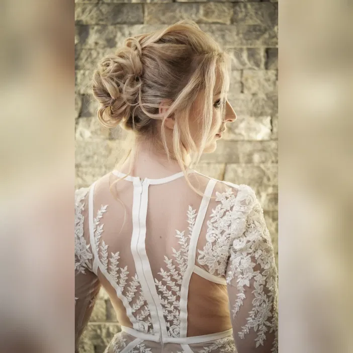 Zbliżenie na tył sukni ślubnej z koronkowymi zdobieniami i eleganckim upięciem włosów na tle kamiennej ściany.