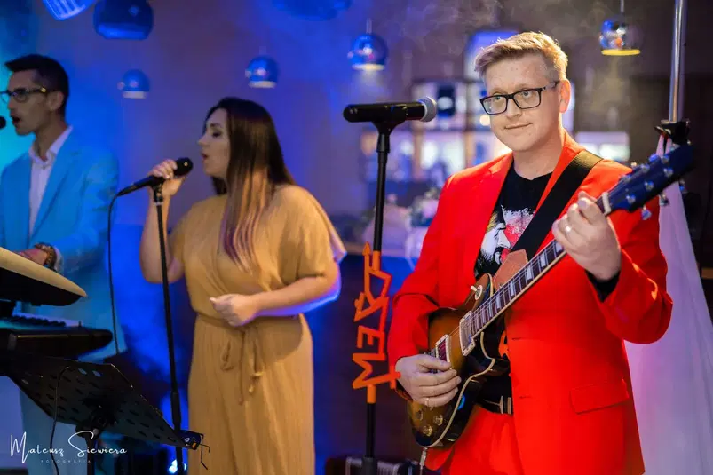  Zespół muzyczny na scenie podczas występu, mężczyzna w czerwonym garniturze gra na gitarze, kobieta śpiewa, mężczyzna w błękitnym garniturze gra na keyboardzie. 