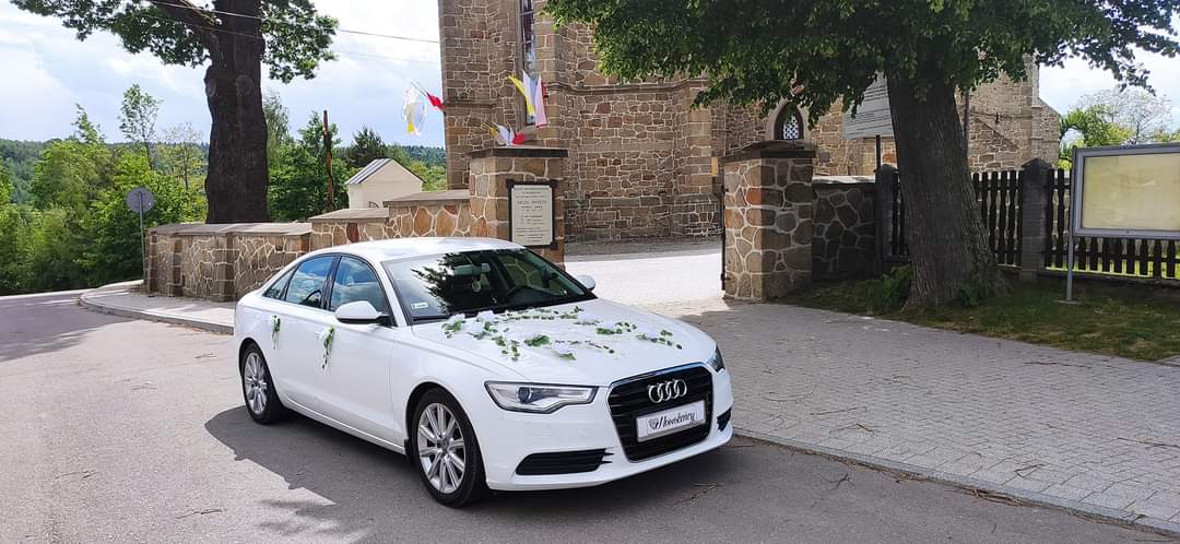  Białe Audi ozdobione kwiatami na tle kamiennego kościoła w słoneczny dzień. Samochód ślubny przed kościołem. 