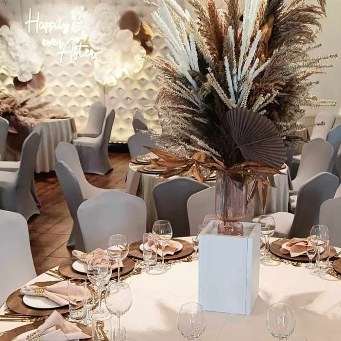 Elegancka sala weselna z dekoracjami kwiatowymi, stoły bankietowe z zastawą i napis Happily Ever After na ścianie dekoracyjnej.