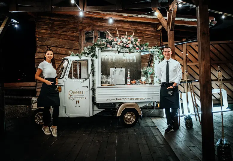  Mobilny bar Prosecco Van podczas eleganckiego eventu w drewnianej altanie, ozdobiony kwiatami i nastrojowym oświetleniem. 