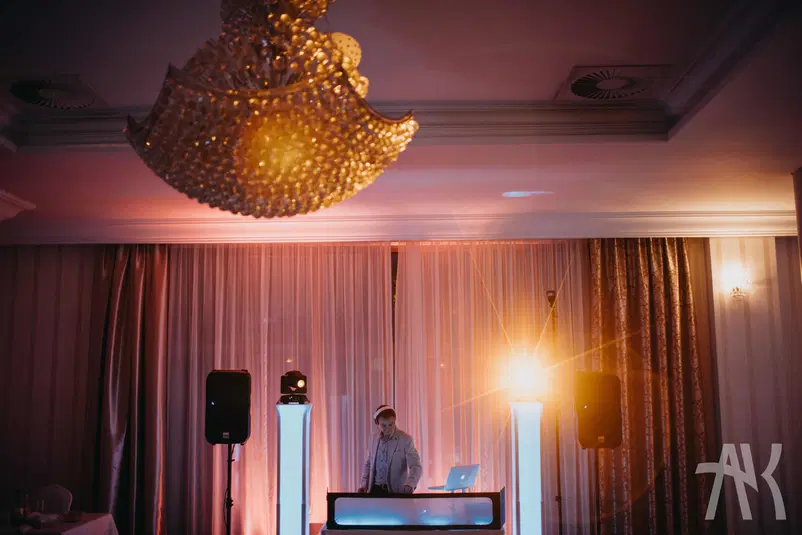  DJ na scenie w eleganckiej sali, efektowne oświetlenie, kryształowy żyrandol, muzyczna atmosfera na wydarzeniu. 