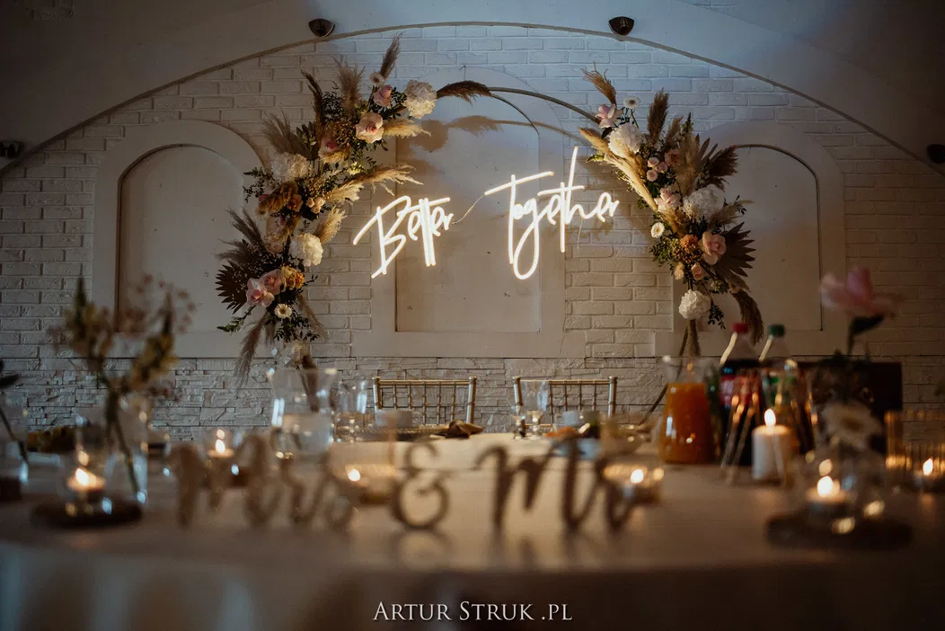 Dekoracja weselna z neonowym napisem Better Together na tle białej cegły, ozdobiona kwiatowymi aranżacjami z suszonymi roślinami, świecami i eleganckim stołem w restauracji.