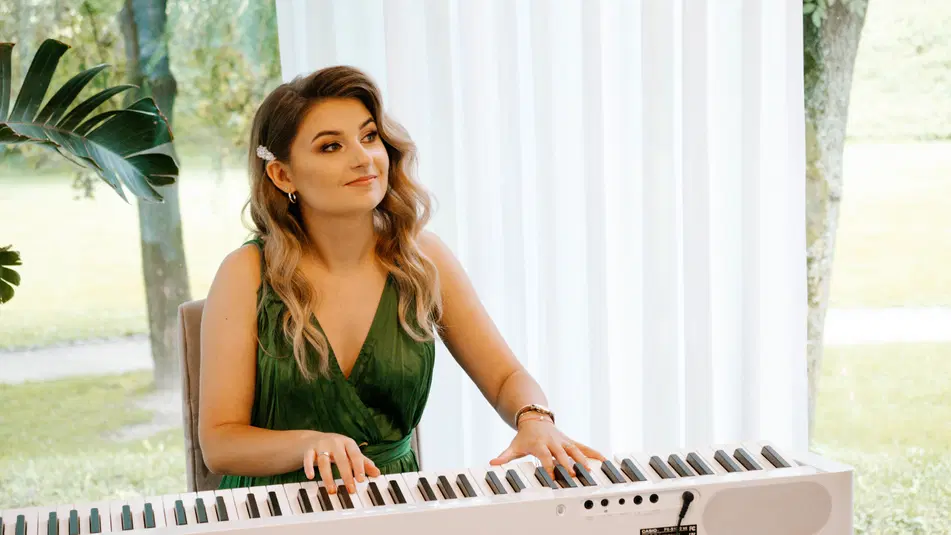  Kobieta w zielonej sukience grająca na keyboardzie, wnętrze z dużym oknem w tle. 