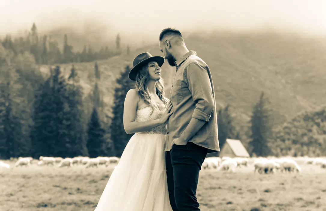 Para w romantycznej scenerii górskiej, kobieta w sukni i kapeluszu, mężczyzna w koszuli, pogodne owce w tle, sielankowy krajobraz, sepia.