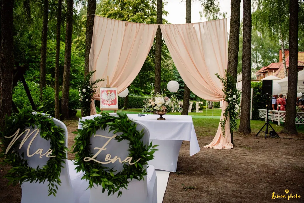 Plenerowa dekoracja ślubna z białymi krzesłami ozdobionymi zielonymi girlandami i napisami „Mąż” i „Żona”, pastelowy baldachim, kwiatowa aranżacja - styl elegancki.