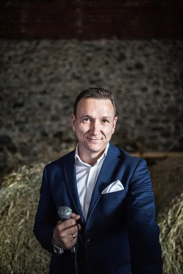  Mężczyzna w garniturze trzymający mikrofon na tle kamiennej ściany i siana. 