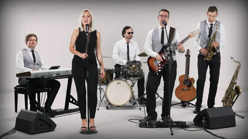  Zespół muzyczny występujący na scenie, pięcioosobowy skład: wokalistka, gitarzysta, perkusista, klawiszowiec i saksofonista w eleganckich strojach i z instrumentami. 