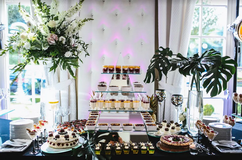 Luksusowy bufet deserowy na przyjęciu weselnym, z eleganckim układem ciast, ciastek i panna cotty, udekorowany świeżymi kwiatami i liśćmi monstery.