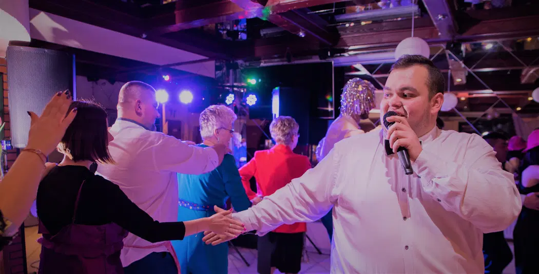  DJ prowadzący imprezę taneczną z mikrofonem, goście bawiący się na parkiecie, kolorowe światła i muzyka na żywo, wesele lub uroczystość rodzinne. 