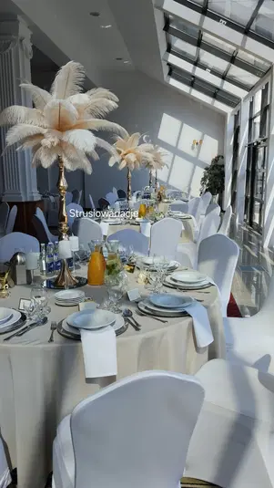 Elegancka dekoracja stołu weselnego z wysokimi wazonami ozdobionymi piórami, białe krzesła z pokrowcami, nowoczesne wnętrze z dużymi oknami. 