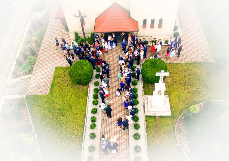  Widok z drona na grupę ludzi przed kościołem, ceremonia ślubna, spotkanie towarzyskie, otoczenie zieleni, chrześcijańska architektura. 