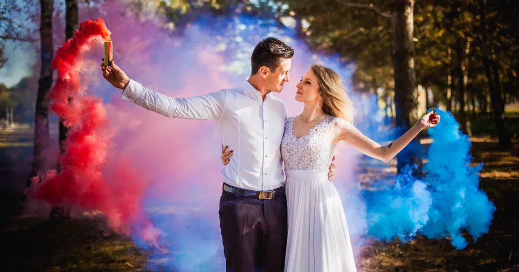  Para trzymająca kolorowe dymy, romantyczna sesja zdjęciowa w plenerze, kobieta w białej sukni, mężczyzna w białej koszuli, atmosfera miłości i harmonii. 