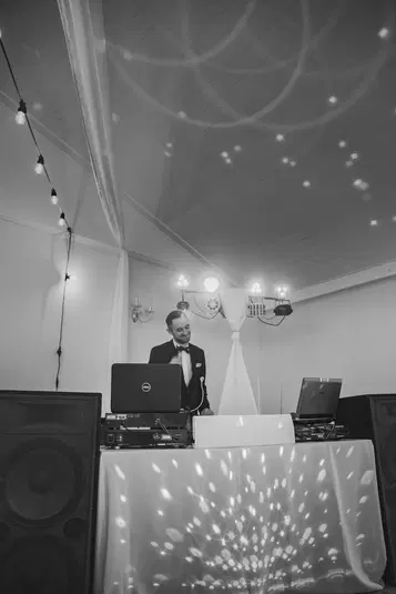  DJ na weselu, scenografia z lampkami, czarno-białe zdjęcie, elegancki mężczyzna przy konsoli, świąteczne światła na suficie, profesjonalna oprawa muzyczna. 