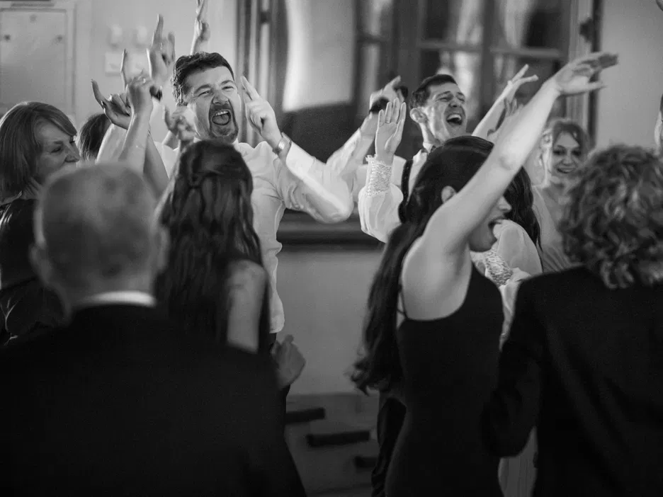 Grupa osób tańczących i świętujących na weselu, czarno-białe zdjęcie z emocjonującymi wyrazami twarzy, wesele, zabawa, radość.