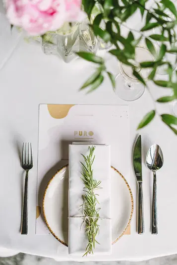  Elegancka aranżacja stołu weselnego z białym obruszem, sztućcami, serwetką z gałązką rozmarynu i kartą menu w tle. 