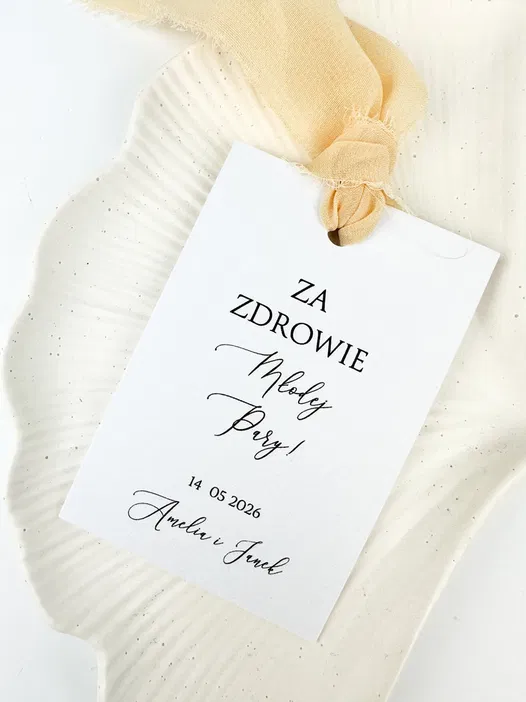 Kartka ślubna z napisem Za zdrowie Młodej Pary! oraz datą 14.05.2026, przewiązana jasną wstążką, leżąca na dekoracyjnym tle.