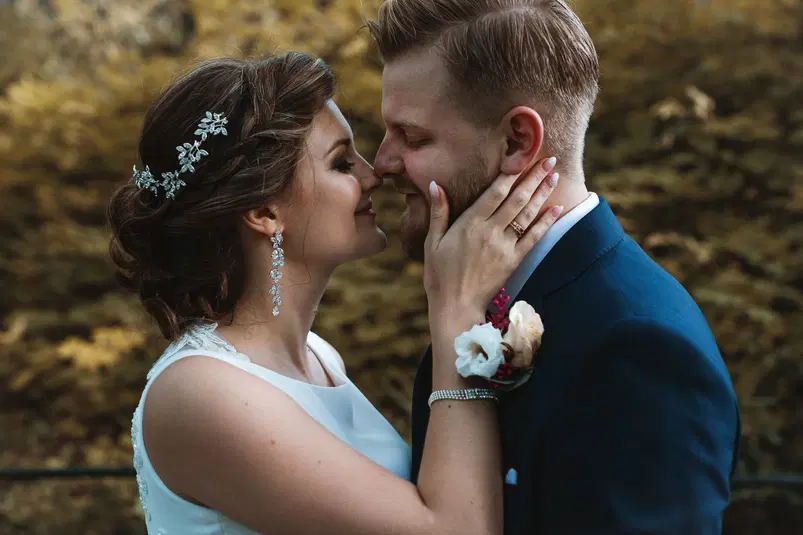  Para młoda w romantycznym uścisku podczas sesji ślubnej w plenerze, elegancka panna młoda w białej sukni i pan młody w granatowym garniturze z kwiatem w butonierce. 