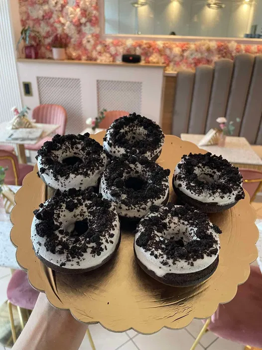 Sześć czekoladowych pączków z białą polewą i kruszonymi ciastkami Oreo na złotej tacy w eleganckiej kawiarni z kwiatową dekoracją.