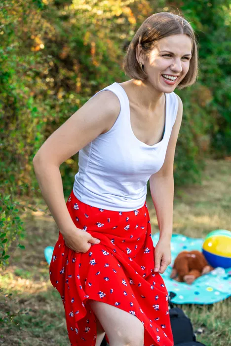 Młoda kobieta w białej bluzce i czerwonej spódnicy na pikniku w słoneczny dzień, uśmiechająca się na tle zielonych krzewów.