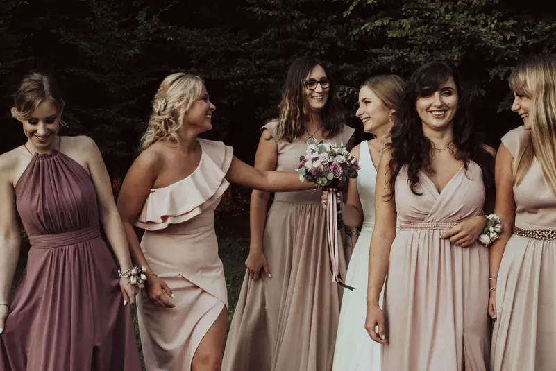  Grupa uśmiechniętych kobiet w eleganckich sukniach, trzymających bukiet kwiatów, stojących na tle zieleni, wesele na świeżym powietrzu. 