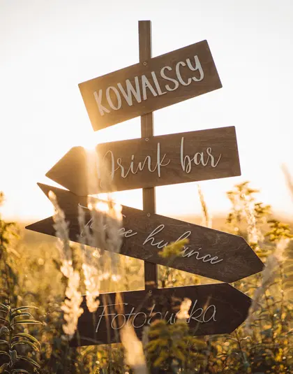 Drewniany drogowskaz weselny na tle zachodzącego słońca z napisami „Kowalscy,” „Drink bar,” „Tańce hulance,” „Fotobudka” w polu traw oświetlonym złotym światłem. 