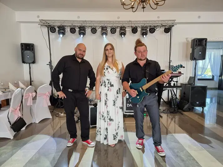  Trio muzyczne przed występem w eleganckiej sali bankietowej, z gitarzystą w czerwonych trampkach i kobietą w białej sukience w kwiaty. 