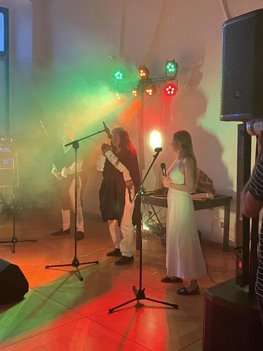 Koncert na żywo, kolorowe światła sceniczne, występ muzyczny z wokalem, ludzie na scenie, muzyka na żywo, atmosfera koncertowa.