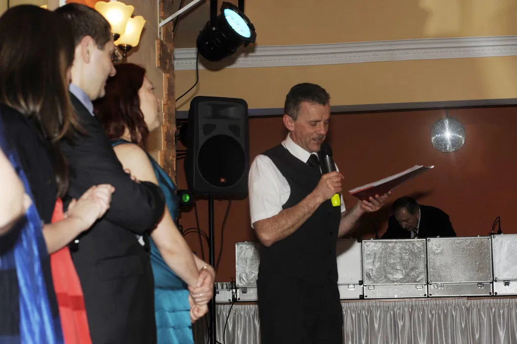Mężczyzna w garniturze przemawia do mikrofonu, stojąc obok grupy ludzi na przyjęciu weselnym w eleganckim wnętrzu z oświetleniem scenicznym.