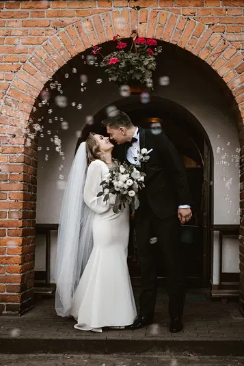  Para młoda stojąca pod łukiem z czerwonej cegły, uśmiechnięta, ubrana w elegancki strój ślubny, w otoczeniu baniek mydlanych. 