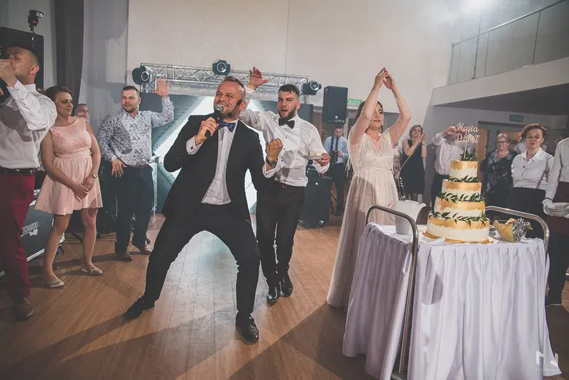  Grupa ludzi bawi się na sali weselnej, z przodu mężczyzna w garniturze śpiewa do mikrofonu, obok widoczny piętrowy tort weselny. 