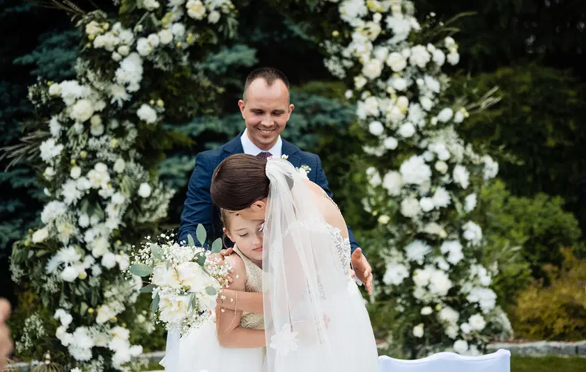  Ślubna ceremonia na świeżym powietrzu, panna młoda w białej sukni ślubnej obejmuje dziecko, kwiatowe dekoracje w tle. 