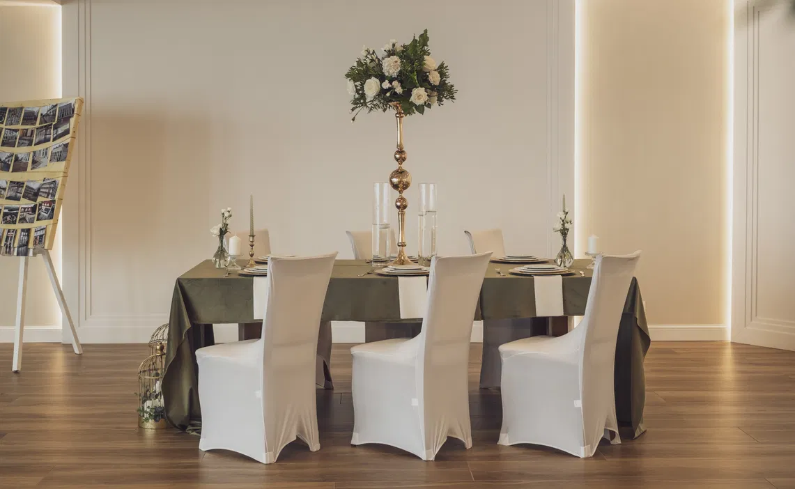 Elegancko nakryty stół na wesele z białymi pokrowcami na krzesła, dekoracją kwiatową i świecami, stylowa aranżacja wnętrza.