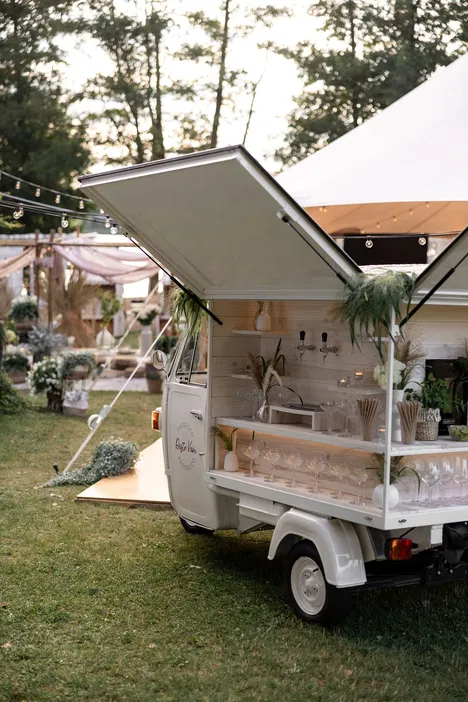 Mobilny bar prosecco na imprezie plenerowej, z elegancką dekoracją i szklankami do serwowania napojów, ustawiony na zielonym trawniku, otoczony dekoracjami ślubnymi i lampkami.