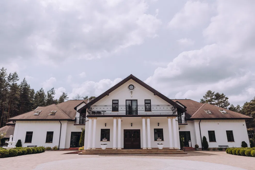 Luksusowa rezydencja z ogrodem na tle lasu, klasyczna architektura, elegancki biały budynek z kolumnami, przestronny podjazd.